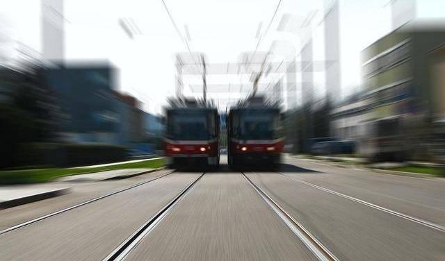 Das Bild zeigt eine Straßenszene mit Langzeitbelichtung. Im Vordergrund sehen wir zwei parallele Bahngleise. Auf den Gleisen befinden sich zwei symmetrische rot-weiße Straßenbahnen.