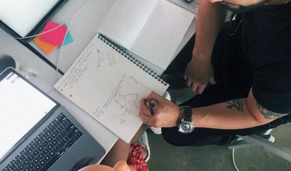 Das Bild zeigt zwei Personen, einen Mann und eine Frau, die an einem Tisch zusammenarbeiten. Der Mann, der einen dunklen Pullover trägt und ein Tattoo auf seinem Arm hat, beugt sich über ein Notizbuch und schreibt etwas hinein. Die Frau, die ein rotes Kleid trägt, sitzt neben ihm und scheint ihm zuzuhören oder ihm zu helfen. Auf dem Tisch liegen Laptops, Notizbücher und verschiedene andere Gegenstände. Die Szene wirkt konzentriert und kollaborativ. Es scheint sich um ein Brainstorming oder Design-Meeting zu handeln, was durch die Notizen im Notizbuch nahegelegt wird. Das Bild vermittelt ein Gefühl der Zusammenarbeit und kreativer Arbeit.