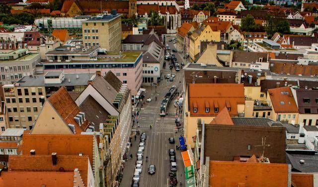 Luftaufnahme in Augsburg einer belebten Straße mit Autos, Bussen und Fußgängern, umgeben von mehrstöckigen Gebäuden und einer Kirche im Hintergrund.