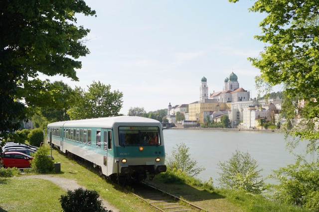 Ein Triebwagen steht am Donauufer auf einer Wiese, in der Mitte fließt die Donau entlang und auf dem gegenüberliegenden Ufer ist die Altstadt Passaus zu erkennen.