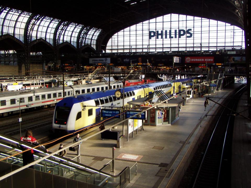Das Foto zeigt die Innenansicht der Bahnhofshalle des Hamburger Hauptbahnhofs. Im Vordergrund befindet sich eine Rolltreppe mit Menschen, die nach oben fahren. Mehrere Gleise verlaufen parallel zueinander, auf denen Züge stehen. Ein Zug in den Farben Blau und Gelb steht im Fokus. Im Hintergrund sind Werbeschilder und Logos verschiedener Unternehmen wie "Philips" und "Burger King" zu sehen. Das Bahnhofsdach ist hoch und gewölbt, mit vielen Fenstern, die Tageslicht hereinlassen. Die Szene vermittelt eine geschäftige und dynamische Atmosphäre. Foto: W. Jansen, pixelio.de 