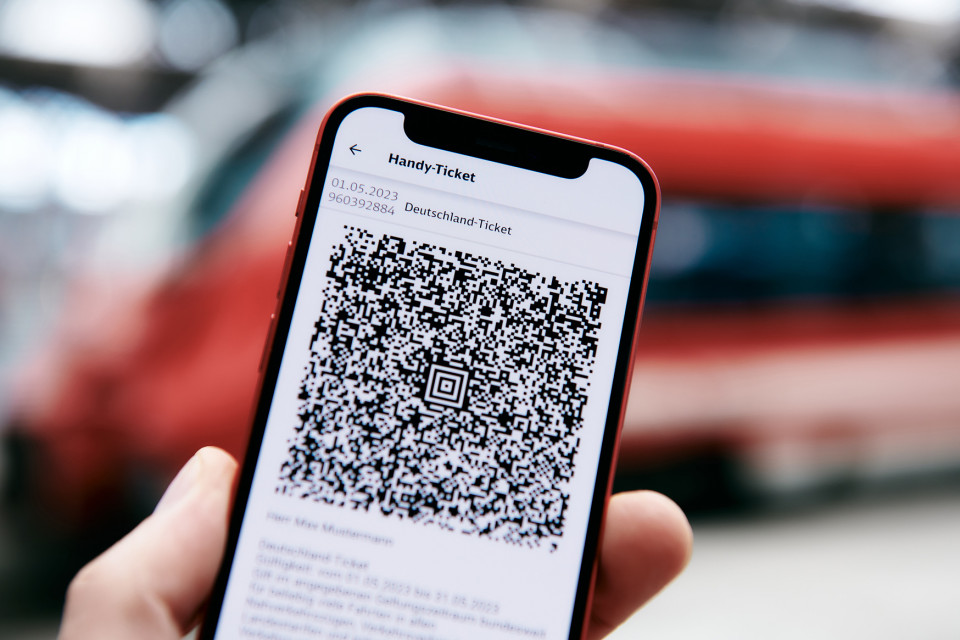 Vor einem ausfahrenden Regionalzug wird ein Handy mit dem Startbildschirm für das 49 Euro-Ticket gehalten.