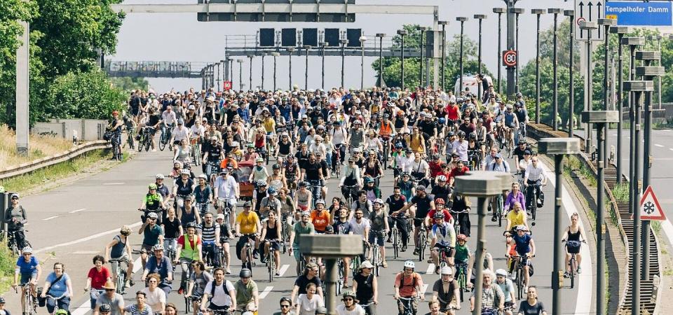 Große Menschenmenge auf Fahrrädern fährt auf einer breiten Straße bei sonnigem Wetter, Verkehrsschilder und Bäume am Straßenrand sichtbar