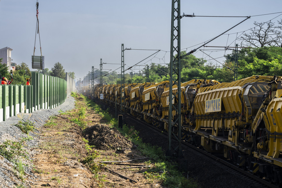 Im rechten Bildteil stehen gelbe Waggons des Umbauzuges im Gleis. Links sind die Fundamente und gesetzte Stützen einer neuen Lärmschutzwand erkennbar. Ein Kran hebt mehrere Segmente der neuen Schallschutzwand ins Bild.