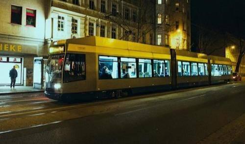 Dunkelblau-silberne Straßenbahn in Leipzig mit eingeschalteten Innenlichtern fährt nachts an beleuchteten Altbauten vorbei.