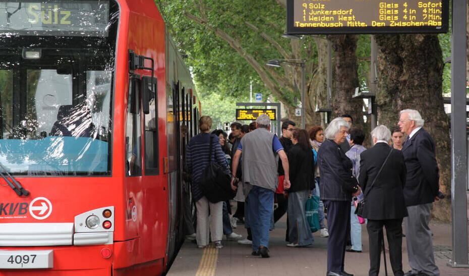 Rote Straßenbahn der KVB mit Fahrgästen an einer Haltestelle unter Bäumen und digitaler Anzeigetafel