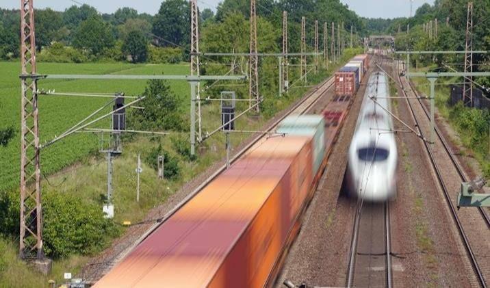 Zwei parallel fahrende Züge auf elektrifizierten Gleisen, ein Güterzug und ein Hochgeschwindigkeitszug, umgeben von grüner Landschaft. Rechts ein ICE, links ein Güterzug.