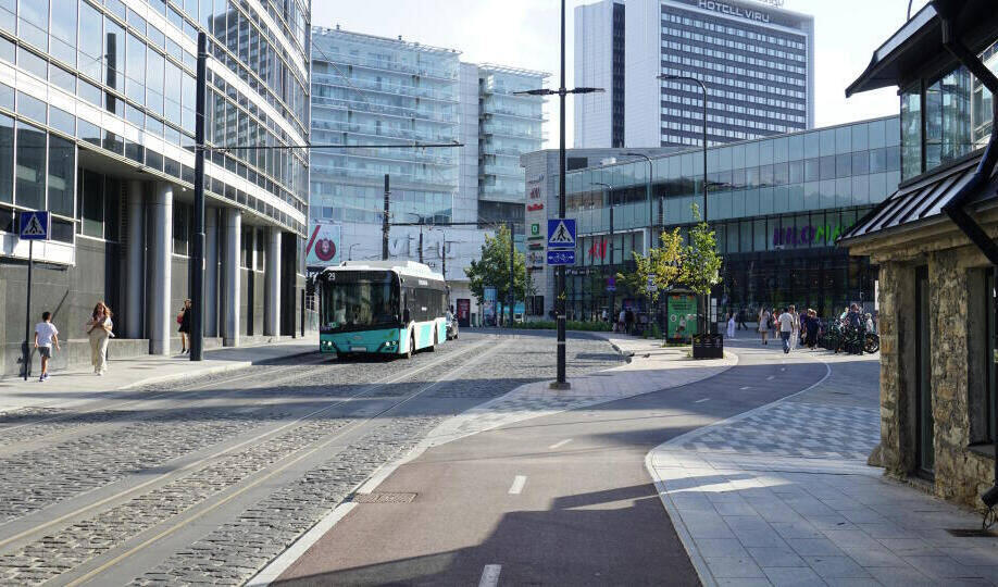 Das Bild zeigt eine moderne, sonnige Straßenszene in einer Stadt. Im Vordergrund verlaufen Straßenbahnschienen auf einer Kopfsteinpflasterstraße. Ein türkisfarbener Bus fährt auf der Straße. Links und rechts befinden sich moderne Gebäude mit Glasfassaden, darunter ein Hotel und ein Einkaufszentrum. Menschen gehen auf dem Gehweg, und im Hintergrund sind weitere Hochhäuser zu sehen.