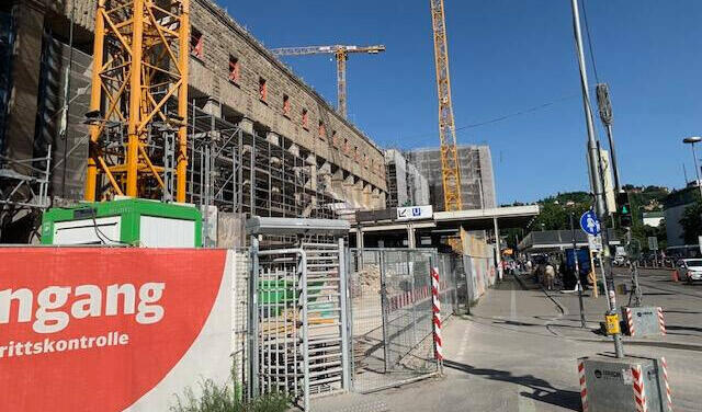 Foto: Klaus Honold Man sieht eine große Baustelle mit mehreren Kränen und Gerüsten. Im Vordergrund befindet sich ein rotes Banner mit den Aufschriften „Eingang“ und „Zutrittskontrolle“ sowie ein Drehkreuz.