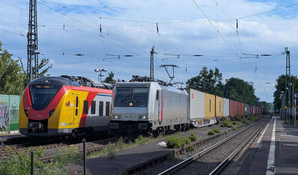 Personenzug und Güterzug nebeneinander auf Gleisen unter Oberleitungen an einem Bahnhof mit Bäumen im Hintergrund