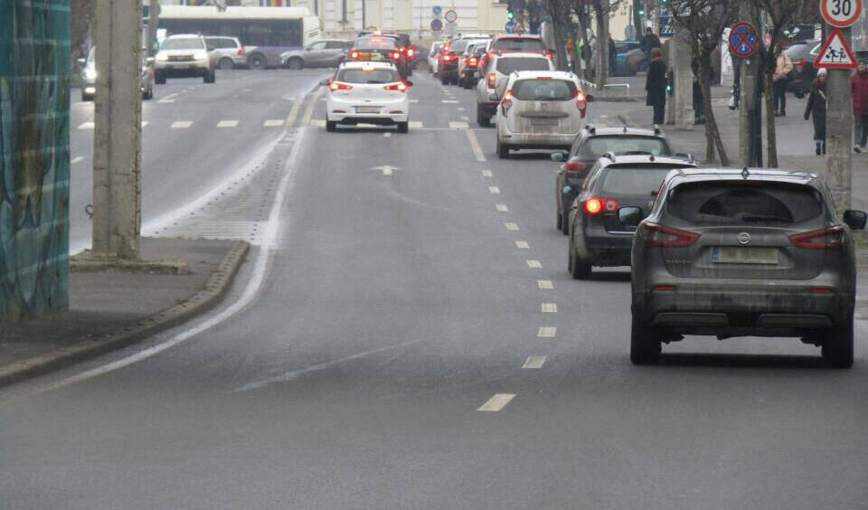 Das Bild zeigt eine belebte Straße in einer europäischen Stadt (erkennbar an den Verkehrszeichen und der Architektur) mit mehreren Fahrspuren und moderatem Verkehr. Autos fahren in beide Richtungen, auf der rechten Seite sind geparkte Fahrzeuge zu sehen. Die Geschwindigkeitsbegrenzung beträgt 30 km/h, und es gibt einen Fußgängerübergang. Im Hintergrund sind historische Gebäude und ein Bus sichtbar, die Ampeln zeigen Grün. Die Straßen sind nass, was auf kürzlichen Regen oder feuchtes Wetter hindeutet. Fußgänger gehen auf dem Bürgersteig, und kahle Bäume deuten auf Herbst oder Winter hin.