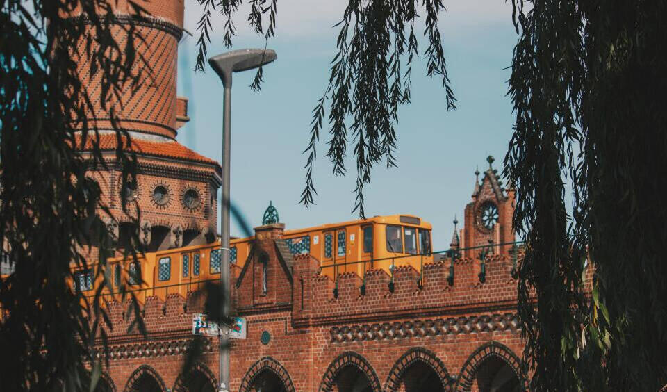 Das Bild zeigt die historische Oberbaumbrücke in Berlin, erkennbar an den markanten roten Backsteinbögen und den beiden Türmen im Stil der norddeutschen Backsteingotik. Eine gelbe Berliner U-Bahn (vermutlich die Linie U1) fährt über die Brücke. Im Vordergrund rahmen hängende Weidenzweige die Szene ein, und eine Straßenlaterne ist sichtbar. Der klare Himmel und das warme Licht verleihen der Aufnahme eine nostalgische, fast malerische Atmosphäre. Die Kombination aus historischer Architektur und typischem Berliner Nahverkehr macht das Bild zu einem charakteristischen Stadtmotiv.