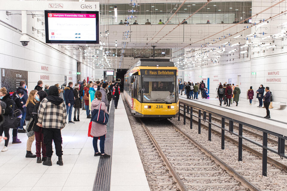 Menschen stehen auf einem Bahnsteig in einer modernen U-Bahn-Station neben einer einfahrenden gelben U-Bahn mit der Anzeige 'E Haus Beuelheim'