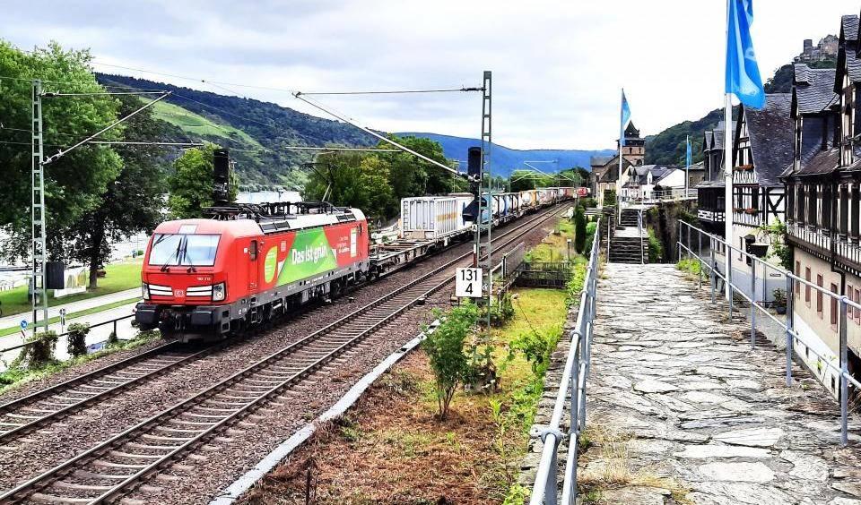 Ein Güterzug mit Containern fährt durch Oberwesell. Links zwischen Bäumen ist der Rhein zu sehen, rechts stehen neben einem Fußweg Fachwerkhäuser