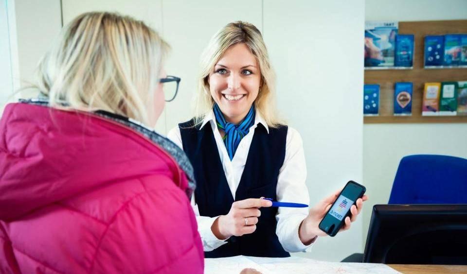 Beraterin zeigt Kundin auf Smartphone das Deutschlandticket mit Logo und erklärt Details am Schalter