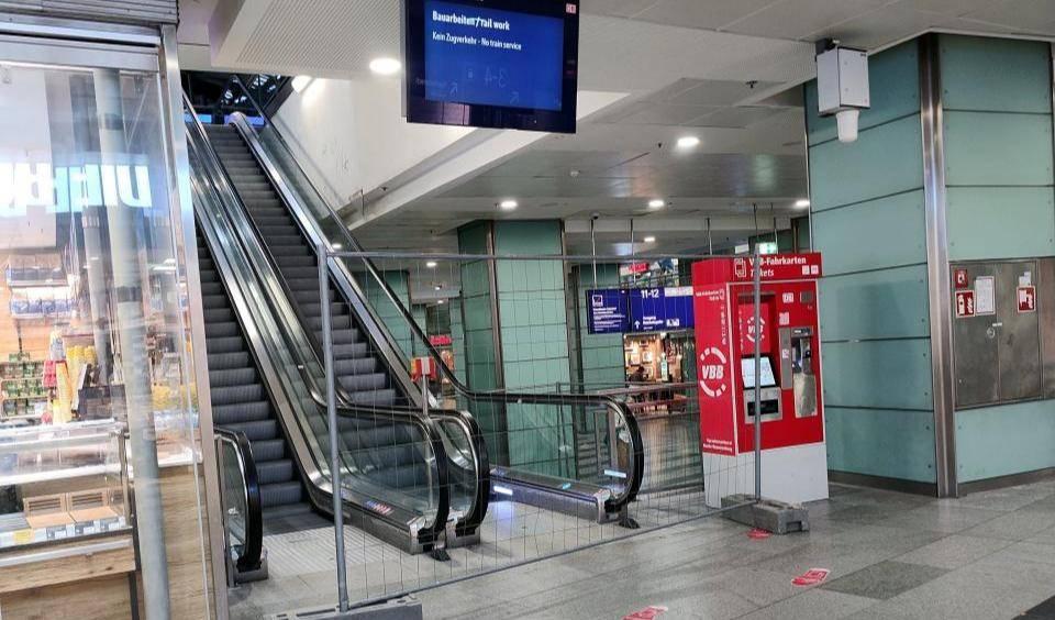 Rolltreppe im Bahnhof Friedrichstraße zum gesperrten Regionalbahnsteig mit Bauzaun, rotem VBB-Fahrkartenautomaten und digitaler Anzeige mit Hinweis 'Bauarbeiten / No train service'