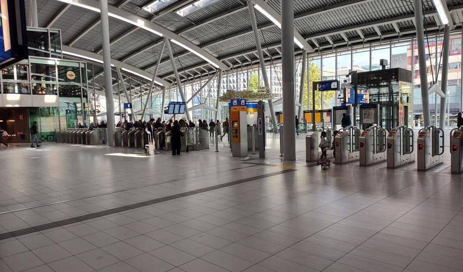 Halle des Bahnhofs Amsterdam Centraal mit hohen Stahlträgern, Fahrkartenautomaten und mehreren Drehkreuzen, Menschen stehen in der Nähe. Soll so auch bald Berlin aussehen?