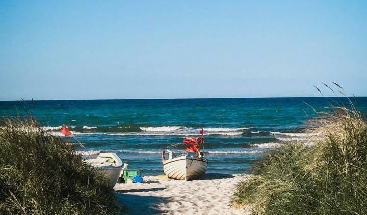 Sandiger Weg zwischen Dünen mit Gras zu einem Strand an der See mit zwei kleinen Booten und ruhiger See unter blauem Himmel.