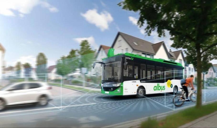 Autonomer Bus 'Albus' von Karsan in grün-weißer Farbgebung, fährt auf einer Straße neben einem Auto und einem Radfahrer. Wohnhäuser im Hintergrund.