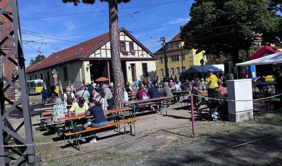 Menschen sitzen an Holzbänken im Freien vor Fachwerkhäusern unter blauem Himmel, einige Bäume und Zelte im Hintergrund.