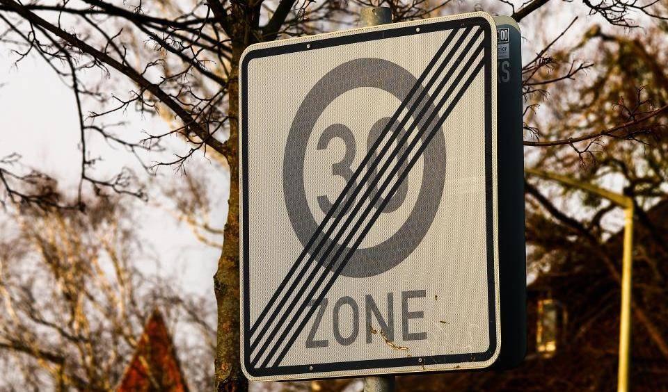 Verkehrsschild mit der Aufschrift '30 ZONE' und diagonalen schwarzen Streifen, umgeben von Bäumen.