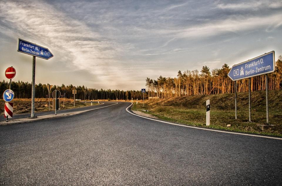   Im Detail zeigt das Bild eine asphaltierte Straße, die sich leicht kurvig durch eine Landschaft schlängelt. Links und rechts der Straße befinden sich Schilder, die auf die Autobahnen nach Frankfurt (O.) und Berlin-Zentrum hinweisen (A10). Ein weiteres Schild zeigt ein Kreisverbot. Zusätzlich sind kleinere Verkehrszeichen und Markierungen auf dem Straßenrand zu sehen. Die Straße ist relativ leer, was auf wenig Verkehr hindeutet. Rechts der Straße erstreckt sich ein Wald aus mittelhohen Bäumen, die im Frühling oder Herbst stehen könnten, da die Blätter eine Mischung aus Braun- und Grüntönen aufweisen. Der Himmel ist leicht bewölkt, mit einer Mischung aus hellen und dunklen Wolken, was eine eher neutrale bis leicht melancholische Atmosphäre erzeugt. Die allgemeine Stimmung des Bildes ist ruhig und friedlich, fast etwas einsam. Die Farben sind gedämpft und eher erdig, mit einem Hauch von warmen Farbtönen im Himmel.